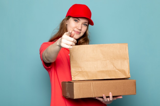 Uma vista frontal feminino atraente correio em camisa polo vermelha boné vermelho segurando pacotes marrons sorrindo posando no trabalho de serviço de alimentação de fundo azul