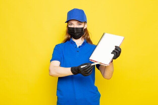 Uma vista frontal feminino atraente courier em camisa polo azul boné azul e calça jeans em luvas pretas enegrecem máscara protetora sobre o trabalho de serviço de alimentação de fundo amarelo