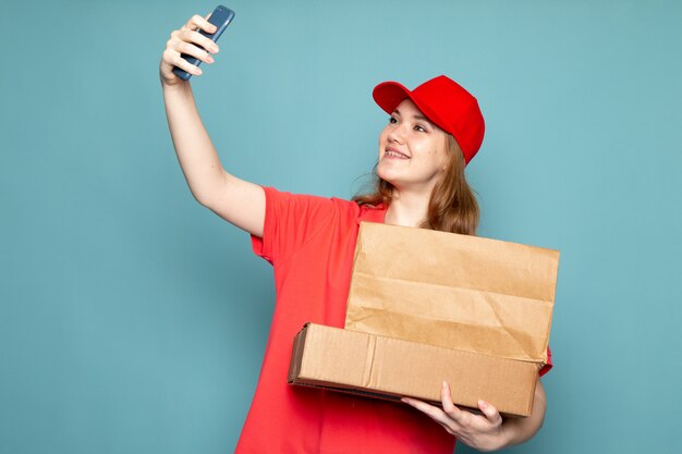 Uma vista frontal feminino atraente courier em camisa polo vermelha boné vermelho segurando o pacote marrom tomando uma selfie no trabalho de serviço de alimentação de fundo azul
