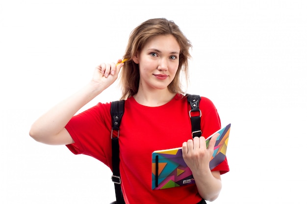 Uma vista frontal jovem aluna na bolsa vermelha camisa preta segurando arquivos de cadernos sorrindo escrevendo para baixo no branco
