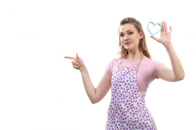 Uma vista frontal jovem atraente dona de casa na capa rosa camisa colorida posando sorrindo segurando pouco coração azul forma sobre o fundo branco cozinha cozinha feminina