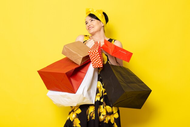 Uma vista frontal jovem atraente em vestido amarelo-preto flor projetado com bandagem amarela na cabeça posando segurando pacotes de compras no amarelo