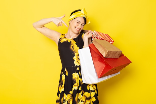 Uma vista frontal jovem atraente em vestido amarelo-preto flor projetado com bandagem amarela na cabeça sorrindo segurando pacotes de compras no amarelo