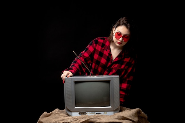 Uma vista frontal jovem bonita camisa quadriculada de vermelho-preto em óculos de sol vermelhos perto de tv pequena