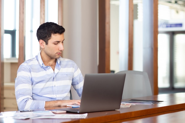 Uma vista frontal jovem bonito na camisa listrada, trabalhando dentro de seu escritório usando seu laptop prateado durante a atividade de trabalho durante o dia