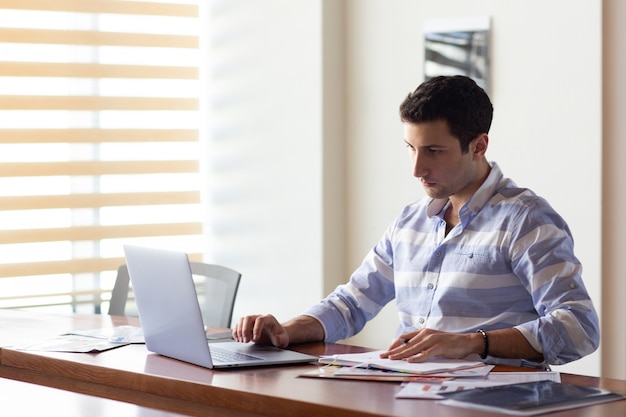 Uma vista frontal jovem bonito na camisa listrada, trabalhando dentro de seu escritório usando seu laptop prateado durante a atividade de trabalho durante o dia