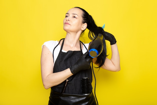 Uma vista frontal jovem cabeleireiro feminino em capa branca de camiseta preta em luvas pretas, segurando o secador de cabelo e escova para secar o cabelo