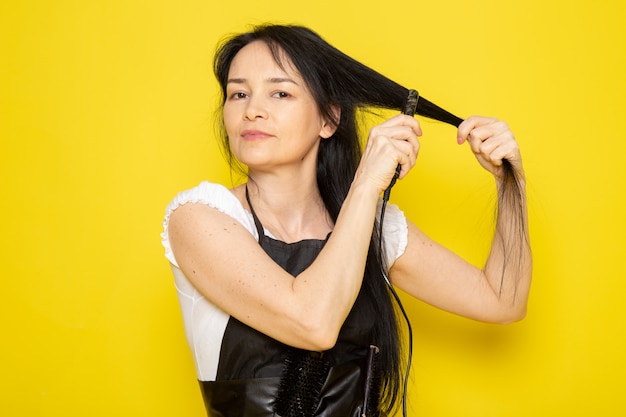 Uma vista frontal jovem cabeleireiro feminino na capa branca de camiseta preta com escovas, ajeitando o cabelo da onda do cabelo