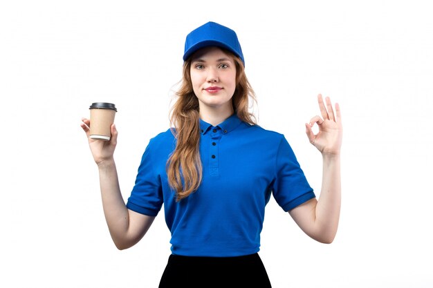 Uma vista frontal jovem correio feminino de camisa azul, boné azul e calça preta, sorrindo, segurando a xícara de café em branco
