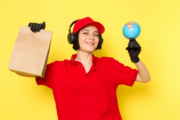 Uma vista frontal jovem correio feminino em luvas pretas uniformes vermelhas e boné vermelho segurando a caixa de alimentos e pouco globo