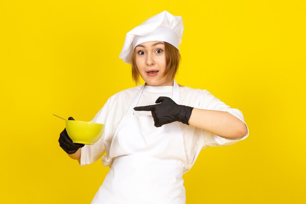 Uma vista frontal jovem cozinheira feminina no fato de cozinheiro branco e boné branco em luvas pretas, segurando a placa verde com espaguete no amarelo