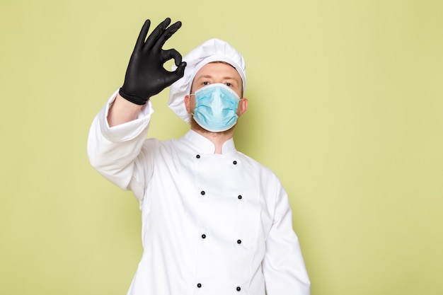 Uma vista frontal jovem cozinheiro masculino em branco cozinheiro terno branco cabeça boné em luvas pretas azul máscara protetora mostrando sinal de tudo bem