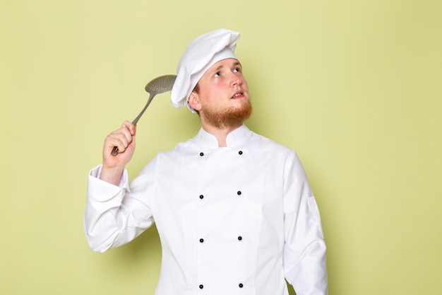 Foto grátis uma vista frontal jovem cozinheiro masculino em branco cozinheiro terno branco cabeça cap segurando grande colher de prata pensando
