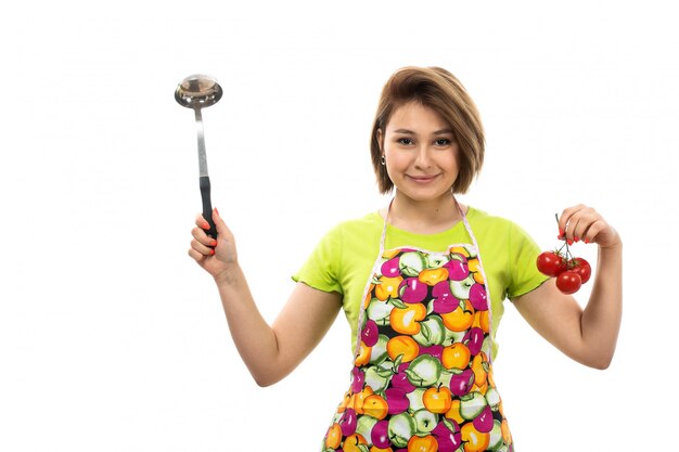 Uma vista frontal jovem dona de casa bonita na capa verde camisa colorida segurando tomates vermelhos e colher de prata sorrindo sobre o fundo branco casa cozinha feminina