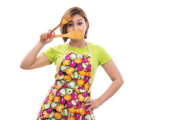 Uma vista frontal jovem dona de casa bonita na capa verde camisa colorida sorrindo segurando o aparelho de cozinha de madeira sobre o fundo branco