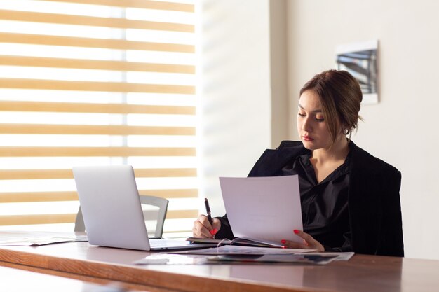 Uma vista frontal jovem empresária atraente na camisa preta jaqueta preta usando seu laptop prata escrita lendo trabalhando dentro de seu escritório trabalho edifício de trabalho