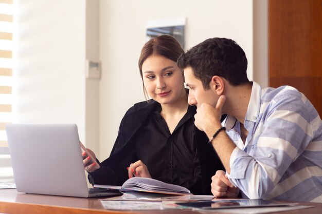 Uma vista frontal jovem empresária linda camisa preta jaqueta preta, juntamente com o jovem usando seu laptop prata discutindo questões dentro de seu escritório trabalho edifício de emprego