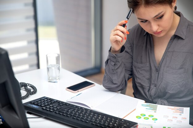 Uma vista frontal jovem empresária trabalhando em seu pc em cima da mesa, juntamente com telefone e gráficos, anotando notas pensando trabalho atividades tecnologia