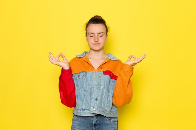 Uma vista frontal jovem fêmea na camisa amarela jaqueta colorida e azul jeans meditando