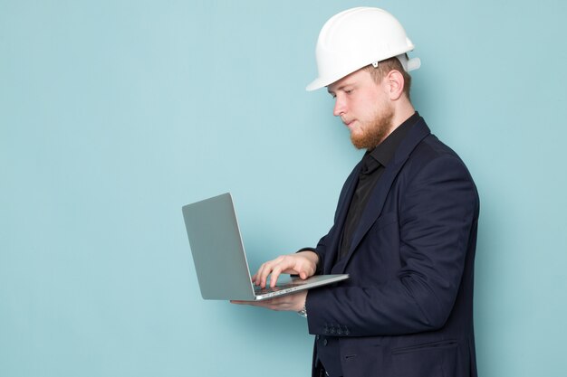 Uma vista frontal jovem homem atraente com barba no capacete de construção preto escuro clássico moderno terno branco usando laptop cinza no espaço azul