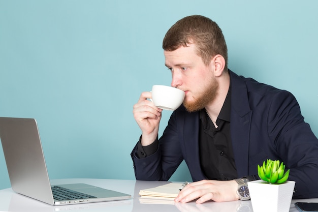 Uma vista frontal jovem homem atraente com barba no terno moderno clássico escuro preto usando laptop tomando café no espaço azul