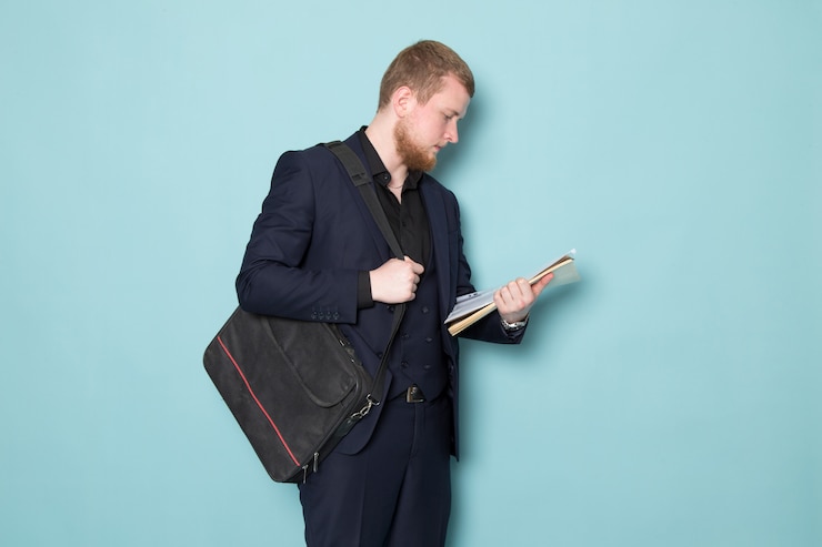 Foto grátis uma vista frontal jovem homem atraente com barba no terno moderno clássico preto escuro, segurando a mochila de arquivos no espaço azul