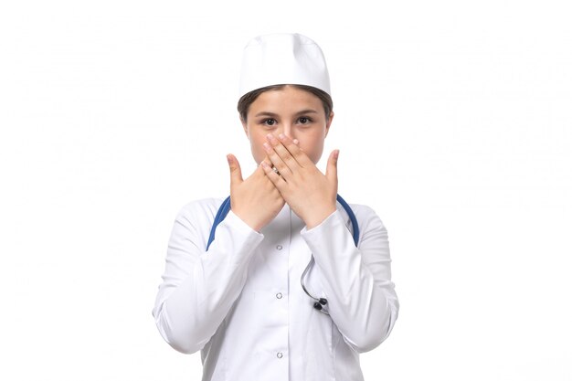 Uma vista frontal jovem médica em traje médico branco com estetoscópio azul posando com expressão tímida