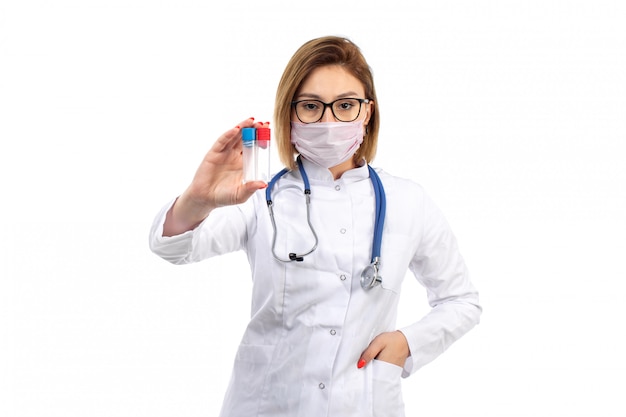 Foto grátis uma vista frontal jovem médica em traje médico branco com estetoscópio usando máscara protetora branca segurando frascos no branco