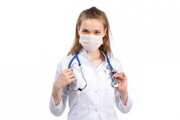 Uma vista frontal jovem médica em traje médico branco com estetoscópio usando máscara protetora branca sobre o branco