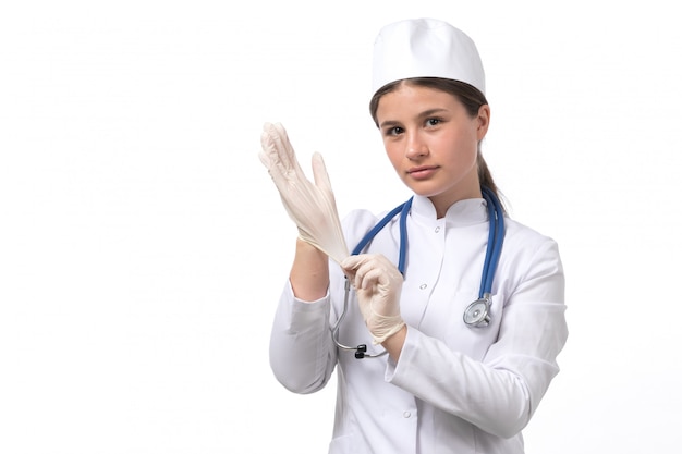 Uma vista frontal jovem médica em traje médico branco e boné branco com estetoscópio azul usando luvas