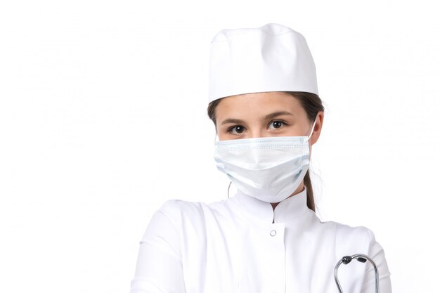 Uma vista frontal jovem médica em traje médico branco e boné branco com estetoscópio azul, vestindo uma máscara