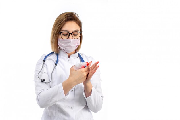 Uma vista frontal jovem médica no terno médico branco com estetoscópio usando máscara protetora branca segurando usando spray no branco