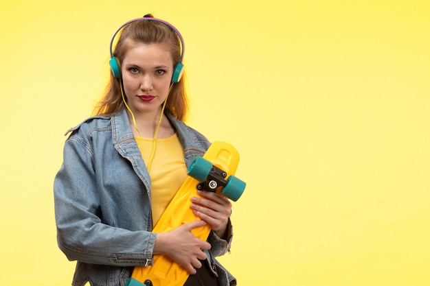 Uma vista frontal jovem moderna de camisa amarela calça preta e jean coat segurando o skate com fones de ouvido coloridos posando