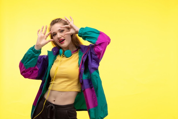 Uma vista frontal jovem mulher moderna em calças de camisa amarela e casaco colorido com fones de ouvido coloridos posando