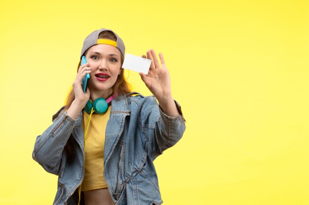 Uma vista frontal jovem mulher moderna em calças de camisa amarela e jaqueta jeans com fones de ouvido coloridos segurando um cartão branco falando ao telefone posando