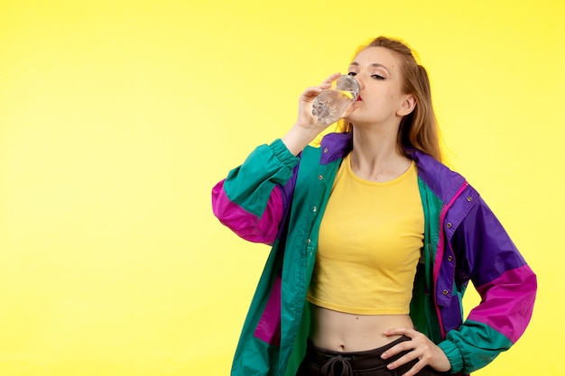 Uma vista frontal jovem mulher moderna em camisa amarela calça preta e jaqueta colorida água potável