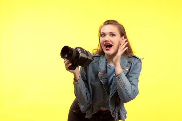 Uma vista frontal jovem mulher moderna em camisa azul calça preta e casaco jean posando surpresa expressão segurando a câmera fotográfica