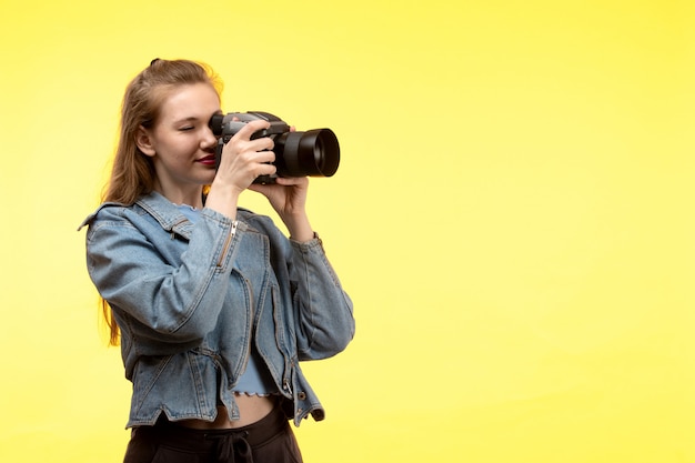 Uma vista frontal jovem mulher moderna em camisa azul calça preta e jean jacket posando expressão feliz sorrindo segurando a câmera fotográfica tirando fotos