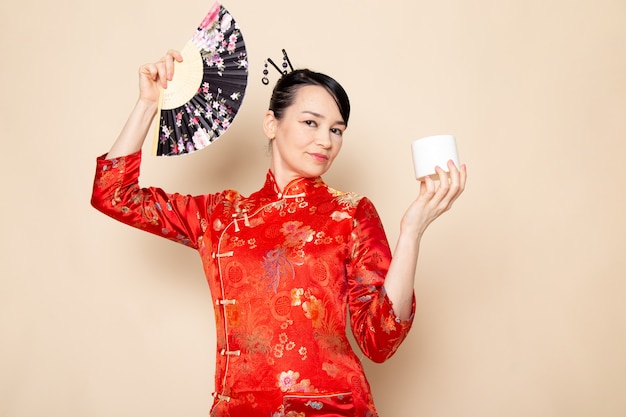 Uma vista frontal linda gueixa japonesa no tradicional vestido japonês vermelho com varas de cabelo posando segurando ventilador dobrável e creme elegante sobre a cerimônia de creme de fundo japão