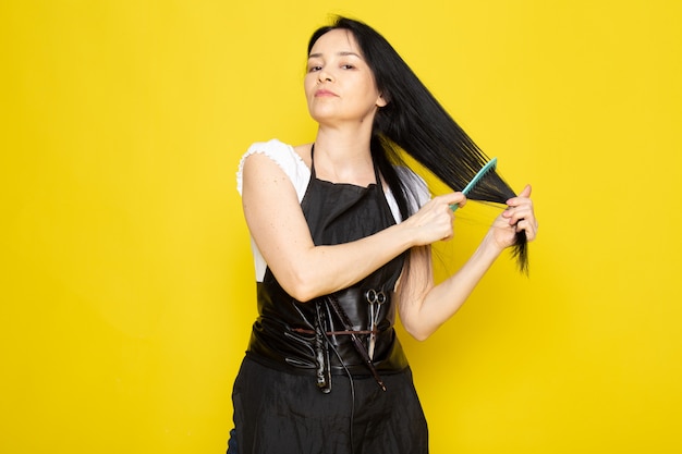 Uma vista frontal lindo cabeleireiro feminino na capa branca de camiseta preta com escovas com cabelo lavado, escovar o cabelo dela posando sobre o cabelo amarelo estilista de fundo amarelo