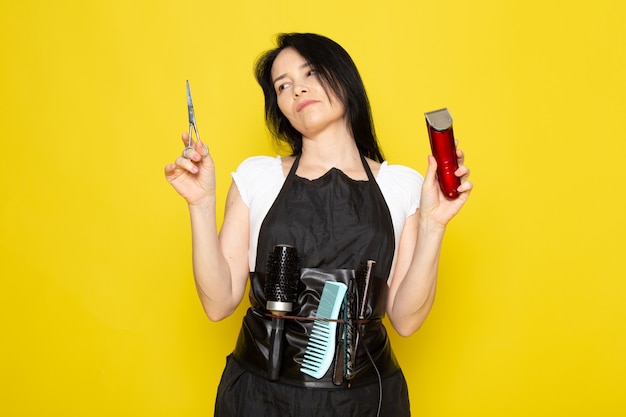 Uma vista frontal lindo cabeleireiro feminino na capa branca de camiseta preta com escovas com cabelos lavados, segurando uma tesoura e uma máquina posando no cabeleireiro estilista de fundo amarelo