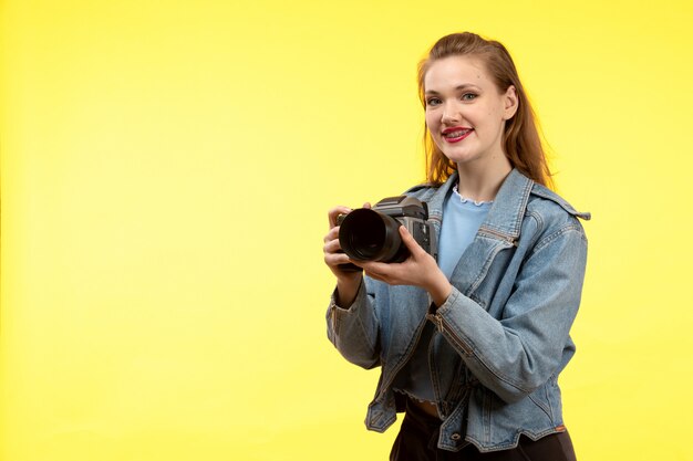 Uma vista frontal mulher moderna jovem de camisa azul calça preta e jean jacket posando expressão feliz sorrindo segurando a câmera fotográfica