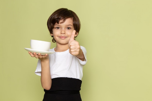 Uma vista frontal, sorrindo, cute, menino, segurando, branca, xícara café, em, branca, t-shirt, ligado, a, pedra colorida, escrivaninha