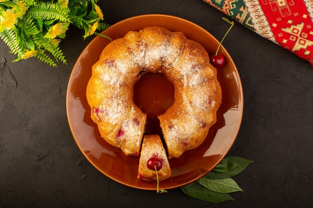 Uma vista superior assada de bolo de frutas delicioso redondo com cerejas vermelhas dentro e açúcar em pó dentro de um prato redondo marrom escuro