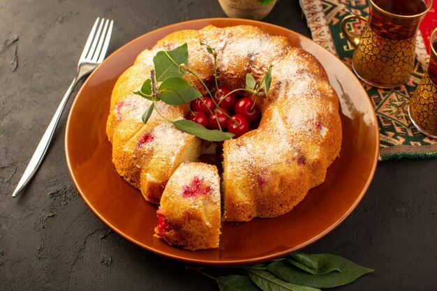 Uma vista superior assada de bolo de frutas delicioso redondo com cerejas vermelhas dentro e açúcar em pó dentro de um prato redondo marrom escuro