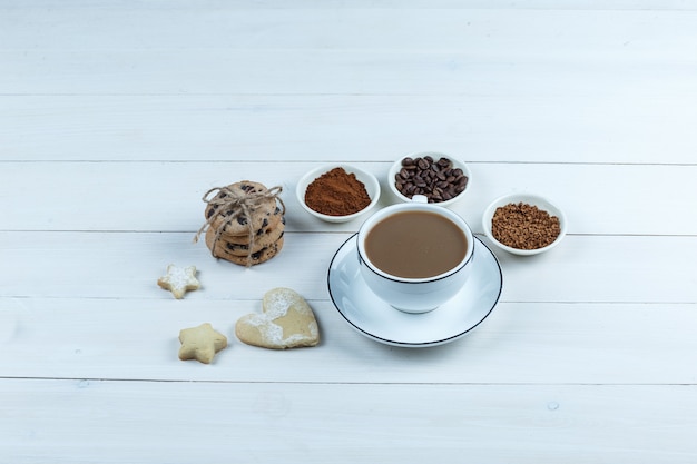 Uma xícara de café com grãos de café, café instantâneo, cacau, diferentes tipos de cookies no fundo da placa de madeira branca, vista de alto ângulo.