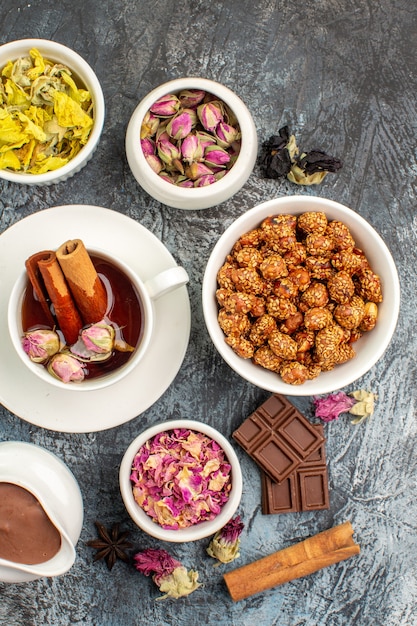 Uma xícara de chá de ervas com diferentes flores secas e chocolate e uma tigela de nozes em solo cinza