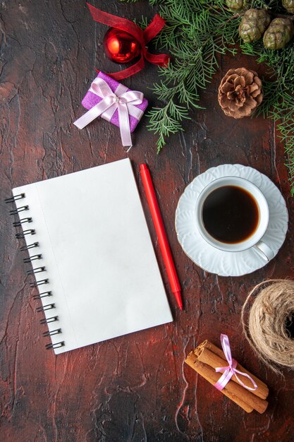 Uma xícara de chá preto, acessórios de decoração de ramos de abeto e um presente ao lado de um caderno com uma caneta em fundo escuro