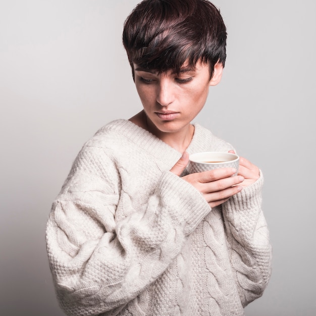Unwell jovem vestindo camisola segurando xícara de café contra um fundo cinza