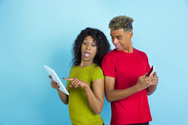 Usando gadgets, rindo, apontando. Jovem emocional afro-americano e mulher com roupas coloridas na parede azul.